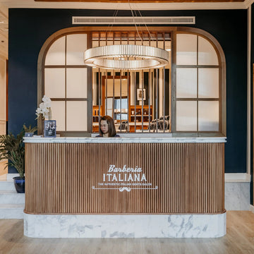 A modern reception desk in a high-end barber shop, featuring a wood slatted front, white marble base, and the text 'Barberia Italiana' on the front. A person is seated behind the counter, with a large arched window behind and wooden shelving on the right.