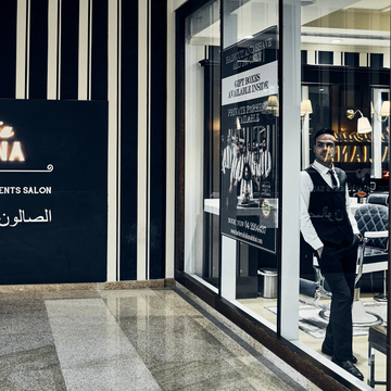 A storefront sign for Barberia Italiana, an authentic Italian hair salon, mounted on a black and white vertically striped wall. The sign features the salon name in elegant gold and white lettering with Arabic text below.