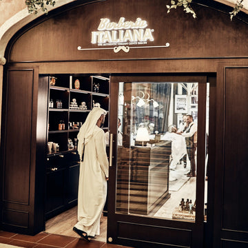 A person in a white traditional robe stands at the entrance of a restaurant or shop with a large arched facade, glass doors, shelves, and a visible sign reading 'Barberia Italiana'.