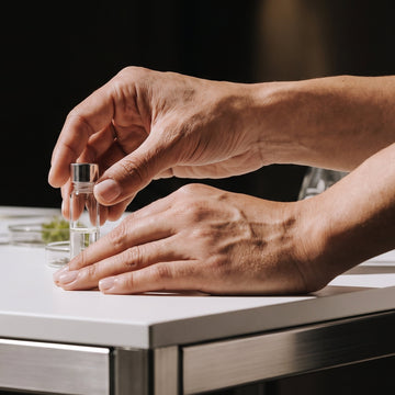 Close-up of hands holding a small glass container on a metallic surface with blurred background